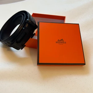 Bälte - Svart snyggt bälte med metal logga och mönster på spännet, kommer i original box och nytt skick