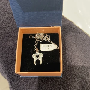 Halsband i silver med tandhänge - Unikt halsband i äkta 925 silver med ett glittrigt tandformat hänge. Hänget är dekorerat med små stenar och har ett hjärtformat hål i mitten. Perfekt för dig som vill sticka ut med ett coolt och annorlunda smycke.