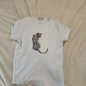Vit t-shirt med leopardtryck från  Subdued - Vit t-shirt från Subdued med ett stort leopardtryck på framsidan. Klassisk rund halsringning och korta ärmar. Mjuk och skön i bomull, perfekt till jeans eller kjol