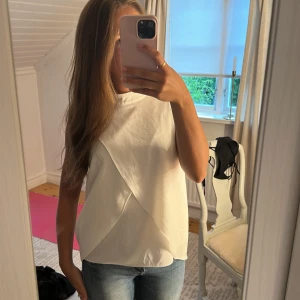 Vit omlott ärmlös blus - Säljer en stilren vit ärmlös blus med snygg omlott-detalj framtill. Toppen har en rak passform och är tillverkad i ett mjukt, lätt material som känns luftigt. Perfekt att matcha med jeans eller kjol för en clean look.