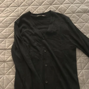 Svart kofta från 157 med knappar - En klassisk svart kofta från 157 i storlek S. Koftan har v-ringning, långa ärmar och stängs med fyra svarta knappar framtill. Perfekt att slänga över en t-shirt eller linne för en chill och stilren look.
