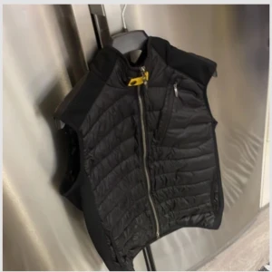 Svart dunväst från Parajumpers M - Säljer en svart dunväst från Parajumpers med quiltad framsida, dragkedja och coolt gult spänne vid halsen. Västen har en bröstficka med dragkedja och en broderad logga på ryggen. Perfekt lager-på-lager-plagg för kyliga dagar.