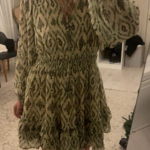 Grönmönstrad kortklänning från Zara - Supersöt kortklänning från Zara med grönt och beige mönster, v-ringning och knappar framtill. Klänningen har långa, luftiga ärmar och smock i midjan för en snygg siluett. Materialet är lätt och flowy, perfekt för vår och sommar.