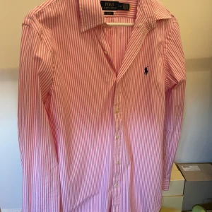 Rosa randig skjorta Polo Ralph Lauren - Säljer en rosa och vit randig skjorta från Polo Ralph Lauren i slim fit. Skjortan har klassisk krage, långa ärmar och knäppning framtill. Broderad blå logga på bröstet. Perfekt för dig som gillar stilrena och fräscha plagg.