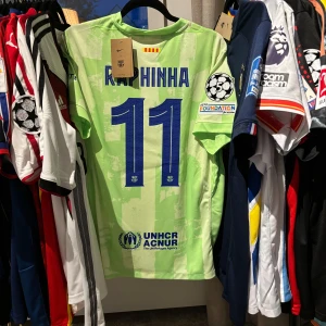 Barcelona Raphinha Champions League fotbollströja - Säljer en Barcelona Champions League-tröja med Raphinha 11 på ryggen. Tröjan är ljusgrön med digitalt camo-mönster, blå detaljer och klubbmärke. Patches från UEFA och UNHCR. Tillverkad i lätt och ventilerande polyester, perfekt för match eller samling. Kolla barcelonamärket