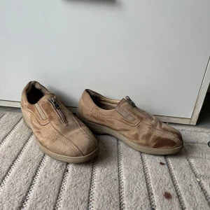 Beige mockaskor med dragkedja från Hästen - Beiga skor från Hästen i mocka med extra bred läst. Skorna har dragkedja framtill istället för snörning och en platt sula. Perfekta för dig som vill ha en enkel och bekväm sko med stilren look.