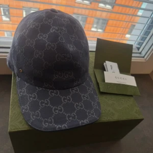 Blå Gucci keps - Snygg blå keps från Gucci med klassiskt GG-monogram i ljusare blått över hela kepsen. Kepsen har böjd skärm och justerbar baksida. Perfekt för dig som vill sticka ut med en lyxig accessoar.