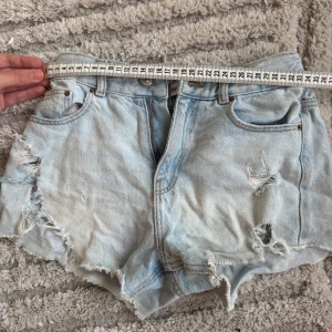 Ljusblå jeansshorts med slitningar - Säljer ett par ljusblå jeansshorts med råa kanter och slitna detaljer framtill. Shortsen har klassisk femficksdesign, knapp och dragkedja samt fransiga benslut för en avslappnad vibe. Perfekta för varma dagar och en cool streetstil.