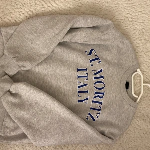 Grå sweatshirt St. Moritz Italy - Säljer en grå sweatshirt med blå text 'ST. MORITZ ITALY' framtill. Tröjan har rund halsringning, långa ärmar och ribbade muddar vid ärmslut och nederkant. Perfekt för en chill och sportig look. finns lite fläckar på nedre armarna som går att få bort