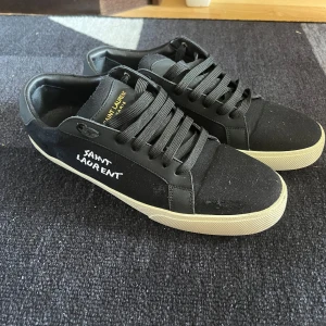 Svarta Saint Laurent sneakers - Svarta sneakers från Saint Laurent med vit logga på sidan och guldtext på plösen. Skorna har svart snörning, rund tå och vit platt sula. Ovandelen är i canvas och detaljer i skinn. Perfekt för dig som gillar stilrena och lyxiga sneakers. OBS orginal skolåda medföljes ej! Använt dessa 5 gånger totalt och dem är för små. STORLEK: 43 men passar bättre på 42. 