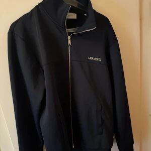  Mörkblå zip tröja från Les Deux - Snygg svart vindjacka från Les Deux med hög krage och dragkedja framtill. Jackan har diskret logga på bröstet och två fickor. Perfekt för dig som gillar stilren och enkel design.
