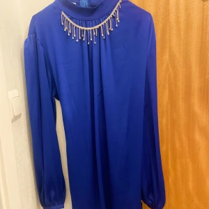 Kornblå långärmad klänning med pärlor - Snygg kornblå klänning med långa ärmar och hög hals. Dekorerad med pärlor och hängande detaljer vid halsringningen som ger en lyxig känsla. Perfekt för dig som vill sticka ut med en elegant och färgstark look.