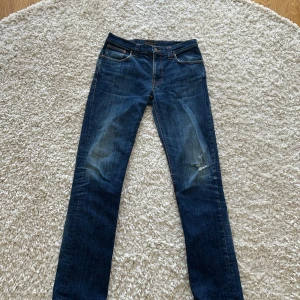 Blå jeans från Nudie Jeans, W34 L36 - Säljer ett par klassiska blå jeans från Nudie Jeans med snygg slitning och orangea sömmar. Modellen har raka ben och fem fickor. Perfekta för dig som gillar en tidlös look med lite edge.