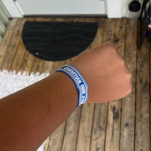 Blått Christian Dior J'Adior armband - Snyggt blått armband från Christian Dior med texten 'J'Adior' broderat i vitt. Armbandet är gjort i tyg och har en justerbar knytning för perfekt passform. Perfekt accessoar för att lyfta din outfit med en lyxig touch.