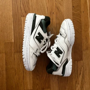 New Balance 550 vita sneakers - Snygga New Balance 550 sneakers i vitt skinn med mörkgröna och svarta detaljer. Klassisk retrodesign med perforerade partier och tydlig logga på sidan. Skorna har snörning och en robust, mönstrad sula för extra grepp. Perfekta för dig som gillar streetstyle.