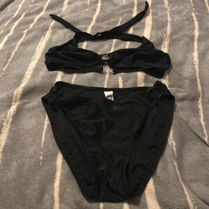 Svart bikini med ringdetalj - Snygg svart bikini från New Wave med halterneck och dekorativ ring i mitten av överdelen. Klassiska bikinitrosor med enkel design. Perfekt för stranden eller poolen, stilren och tidlös look.