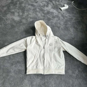 Vit Moncler hoodiejacka med dragkedja - Vit hoodie från Moncler med broderad logga på bröstet. Hoodien har huva med dragsko, två fickor framtill och hel dragkedja. Tillverkad i mjuk bomull för en chill och clean look. Perfekt för dig som gillar stilrena vibes och streetwear.   