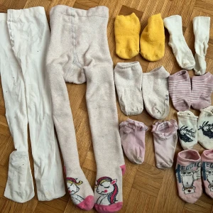 Mix av strumpor och tights i pastell - Flera par strumpor och tights i olika färger och stilar. Inkluderar vita och ljusrosa tights, gula, vita, beiga och rosa strumpor, några med ränder, volang eller tryck med uggla och sjöjungfru. Perfekt för dig som gillar söta och lekfulla detaljer.