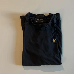 Svart t-shirt från Lyle & Scott - Svart t-shirt från Lyle & Scott med klassisk passform och korta ärmar. T-shirten har det ikoniska gula fågel-loggan broderad på bröstet. Tillverkad i mjuk bomull som känns skön mot huden. Perfekt för en clean och stilren look. Herr strl 