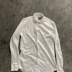 Vit slim fit skjorta från Dressmann - Klassisk vit skjorta från Dressmann i slim fit-modell med extra långa ärmar. Tillverkad i stretchig bomull för en bekväm passform. Skjortan har knappar framtill och vid ärmsluten, perfekt för en clean och stilren look.