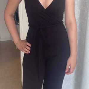 Svart omlott jumpsuit med knytband - Säljer en svart jumpsuit med omlott-topp och djup v-ringning. Byxdelen är rak och något croppad, och midjan markeras med ett knytband. Tyget är ribbat och stretchigt, vilket gör den både bekväm och snygg. Perfekt för dig som gillar stilrena plagg med en twist.