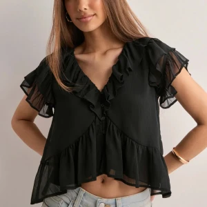 Svart volangblus med v-ringning - Supersöt svart blus med v-ringning och volangdetaljer längs halsringningen och ärmsluten. Blusen är luftig med korta, transparenta ärmar och knappar framtill. Den har en flowy passform och är tillverkad i ett lätt, skirt material som ger en romantisk vibe.
