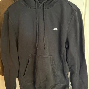 Svart hoodie från J.Lindeberg - Svart hoodie från J.Lindeberg med klassisk passform och liten vit logga på bröstet. Tröjan har huva med snörning och en stor magficka. Tillverkad i mjuk bomull, perfekt för chill dagar eller när du vill ha en clean look. Ganska bra skick