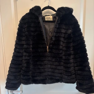 Svart pälsjacka med huva Softy M - Mysig svart pälsjacka från Softy med huva och horisontella ränder. Jackan har dragkedja framtill och är fodrad. Perfekt för kalla dagar när du vill vara både varm och snygg. Materialet är en mix av akryl och polyester.