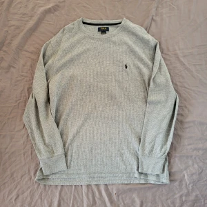 Grå långärmad tröja Polo Ralph Lauren - Grå långärmad tröja från Polo Ralph Lauren i storlek 2XL. Tröjan har ett diskret svart broderat logotyp på bröstet och är tillverkad i ett mjukt, stickat bomullsmaterial med en lätt ribbad struktur. Perfekt för chill dagar och enkel att matcha med jeans.
