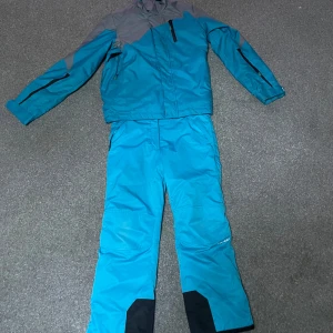 Blå vindjacka från Everest - Blå och grå vindjacka från Everest med dragkedja och hög krage. Jackan har svarta detaljer och praktiska fickor med dragkedja. Materialet är syntet och jackan är perfekt för kalla dagar. Matchande blå byxor med svarta förstärkningar ingår.