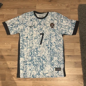 Portugal Ronaldo  - Portugal bortatröja från Nike med Ronaldo och nummer 7 på ryggen. Tröjan är vit med blått abstrakt mönster, svarta detaljer vid ärmslut och krage samt broderat landslagsemblem på bröstet. Tillverkad i lätt och ventilerande material, perfekt för fotbollsälskare.