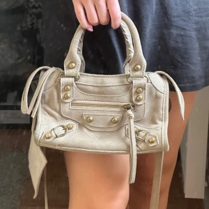 Beige mocka handväska med detaljer - Snygg beige handväska i mocka med guldiga nitar och dragkedja framtill. Väskan har både handtag och axelrem, samt en cool spegel-detalj hängande på sidan. Några fläckar på väskan som jag inte testat att ta bort.