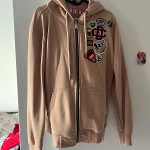 Beige hoodie från Dsquared2 med patches - Säljer en beige hoodie från Dsquared2 med coola broderade patches på bröstet och dragkedja framtill. Tröjan har huva med snörning, kängurufickor och Dsquared2-logga i rött på ryggen. Perfekt för dig som gillar streetwear och snygga detaljer.