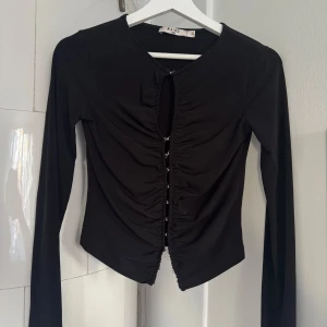 Svart långärmad topp från NA-KD XS - Svart långärmad topp från NA-KD i storlek XS. Toppen har snygg rynkad design framtill med dekorativa silverfärgade hakar som stängning längs hela framsidan. Figurnära passform och mjukt stretchigt material🫶🏼