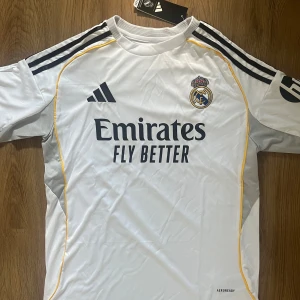 Real Madrid 2025/2026 Hemma Tröja - Real Madrid hemmatröja från Adidas i slim fit. Vit med svarta detaljer och gula inslag längs sidorna. Broderat klubbmärke på bröstet, Adidas-logga och 'Emirates Fly Better' tryck. Tillverkad i lätt och ventilerande AEROREADY-material.                 Real Madrid 2025/2026 Home Kit, Storlek S, aldrig använd med lappen kvar.