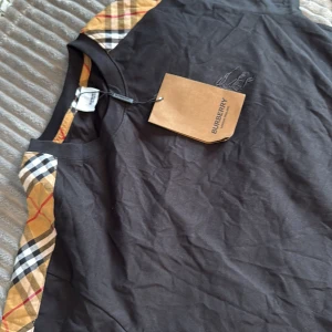 Svart Burberry t-shirt med rutiga detaljer - Svart t-shirt från Burberry med klassiska beige, röda och svarta rutiga paneler på axlarna. Ikonisk broderad logga på bröstet och rund halsringning. Skön passform och tillverkad i mjuk bomull. Perfekt för dig som vill ha en stilren men ändå unik look.