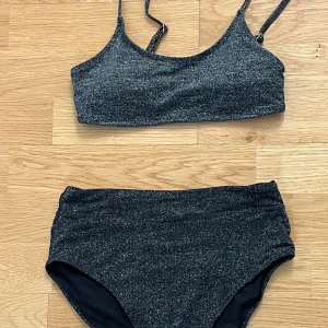 Glittrig svart bikini med hög midja - Supersnygg svart bikini med glittrigt tyg och sportig topp med smala axelband och metallspänne i ryggen. Underdelen har hög midja för en trendig look. Perfekt för dig som vill sticka ut på stranden i sommar!
