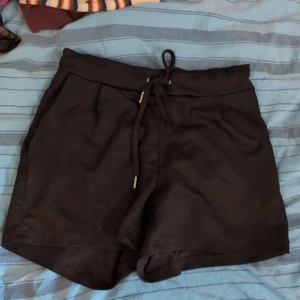Svarta shorts med snörning från Amisu - Svarta shorts från Amisu i mjukt material med elastisk midja och snörning framtill med metalländar. Shortsen har två fickor och en avslappnad passform, perfekta för chill dagar eller träning. Klassisk look som funkar till det mesta.