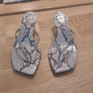 Ormskinnsmönstrade sandaler H&M - Snygga sandaler från H&M med ormskinnsmönster i grått och beige. Skorna har fyrkantig tå, smala remmar och låg klack. Perfekta för dig som vill sticka ut med trendiga detaljer och en cool vibe. Materialet är syntet och de har en stilren look.