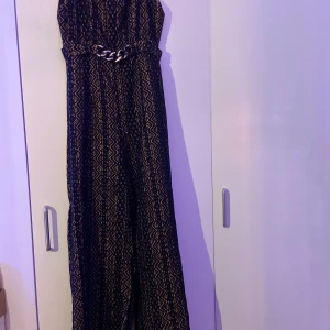 Svart spetsmönstrad byxdress Lady Sunshine - Supersnygg svart byxdress från Lady Sunshine med spetsmönster och nude-färgad underklänning. Vida ben och markerad midja med kedjedetalj. Perfekt för dig som vill sticka ut med coola detaljer och unik look.