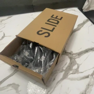 Svarta Adidas Yeezy Slide sandaler - Svarta Adidas Yeezy Slide sandaler i minimalistisk design. Tillverkade i syntetmaterial med öppen tå och platt sula. Perfekta för chill dagar eller när du vill ha en clean och modern look. Kommer med originalkartong och Adidas-påse.