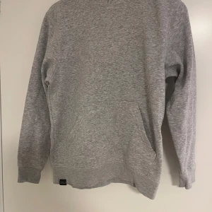 Grå hoodie från South West - En basic grå hoodie från South West med känguruficka och huva. Tröjan är i mjuk bomullsblandning och har långa ärmar samt ribbade muddar. Perfekt för chill dagar eller när du vill ha en enkel och clean look.