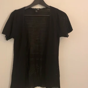 Svart stickad kofta från H&M - Svart  kofta från H&M med korta ärmar och v-ringning. Koftan har fina spetsdetaljer längs halsringningen och stängs framtill med små hyskor. Perfekt att slänga över ett linne eller klänning för en chill look.💗 