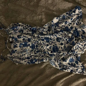 Vit och blå blommig klänning  - Supersöt kortklänning med blått blommönster på vit botten. Klänningen har smala axelband och en lätt, flowy passform som känns perfekt för varma dagar. Materialet är mjukt och svalt, vilket gör den riktigt skön att bära.