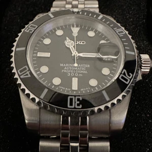 Seiko submariner mod jubilee armband - Säljer en automatisk Seiko submariner mod med svart urtavla, silverfärgat boett, safirglas och jubilee länk i rostfritt stål. Klockan har tydliga index, datumfönster med förstoringsglas och vridbar bezel. Lume på visare och index för extra synlighet. Extra länkbitar och verktyg medföljer. Köpt på LumeLab för 3000kr. Priset kan diskuteras