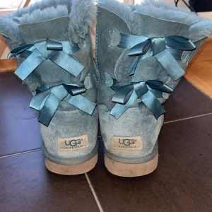 Blå UGG boots med rosetter - Mysiga blå UGG boots i mocka med fluffig insida och dekorativa satinrosetter baktill. Skorna har rund tå och platt sula, perfekta för kalla dagar. Klassisk UGG-logga på hälen och snygg söm längs kanterna. Ganska vintage. 