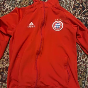 Röd Adidas FC Bayern München jacka  - Säljer en röd Adidas jacka med FC Bayern München-logga på bröstet. Jackan har dragkedja framtill, ståkrage och två fickor. Perfekt för dig som älskar fotboll och vill ha en sportig look. Klassisk Adidas-design med broderad logga och klubbmärke. Strl M men passar perfekt för dig som i normalfallet har strl S