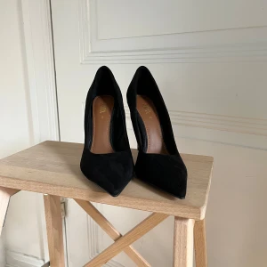 Svarta pumps i mocka från Zara - Stilrena svarta pumps från Zara i mjuk mocka med spetsig tå och hög klack. Snygg siluett och klassisk design som lyfter varje outfit. Klacken har en elegant finish och insidan är i brunt skinnliknande material.