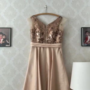 Champagnefärgad långklänning med paljetter - Långklänning i champagnefärg med glittrig paljettöverdel och v-ringning. Kjolen är slät och faller snyggt, medan överdelen har snörning i ryggen och meshdetaljer vid axlarna. Perfekt för bal eller fest, riktigt elegant och unik look.aldrig använd 