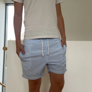 Randiga blå shorts från USPA - Snygga blå och vita randiga shorts från USPA med dragsko i midjan och broderad logga på vänster ben. Sköna fickor på sidorna och klassisk somrig look. Perfekta för chill dagar eller strandhäng.
